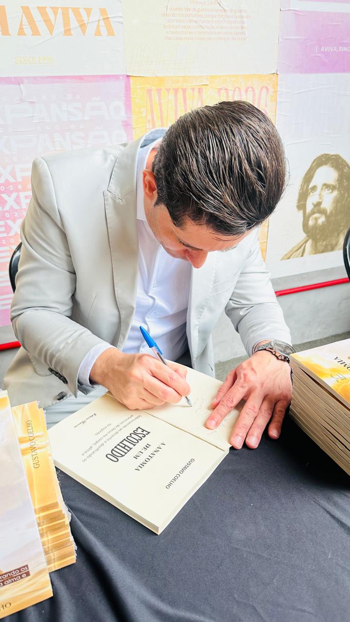 Gustavo Coelho autografando um exemplar de A Anatomia de um Escolhido diante de um painel com a arte Expansão
